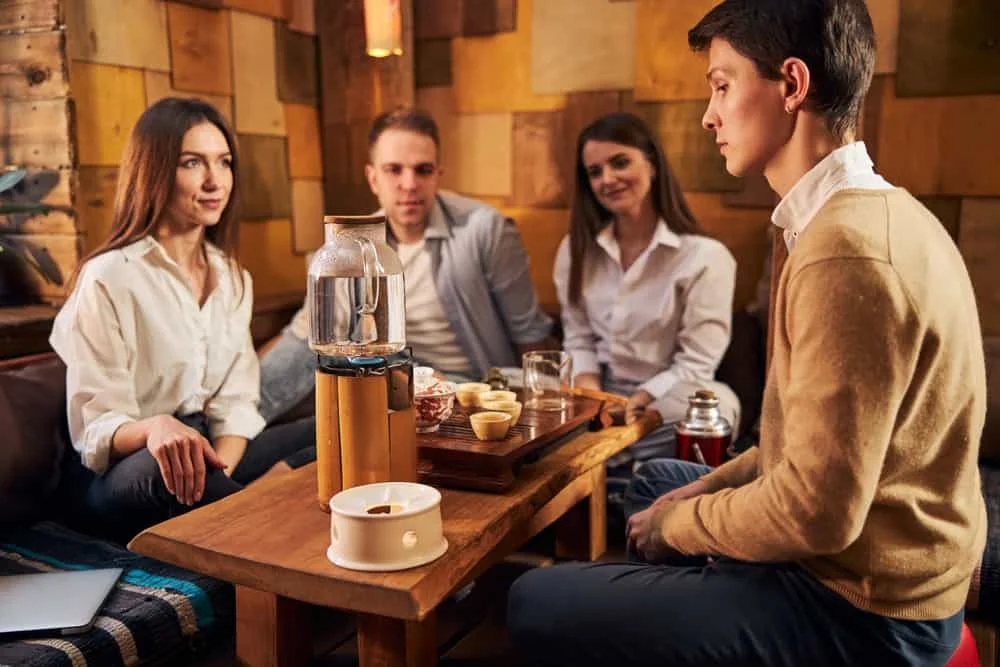 man looking at glass kettle on portable gas burner while sitting at the table with young people in tea house